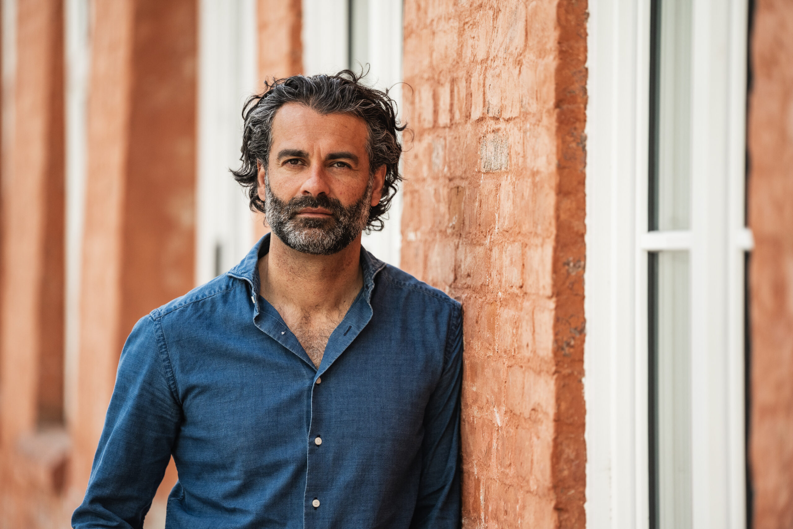 Bilde av Babak Behrad fra Burson AS, står ved en rød murvegg i blå skjorte